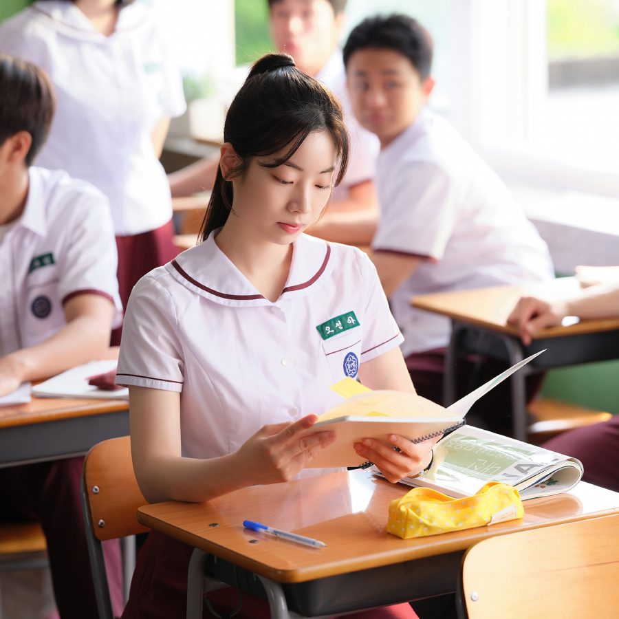 A neatly dressed girl sits reading a book at her desk in a classroom full of students, in a scene from You Are the Apple of My Eye. 