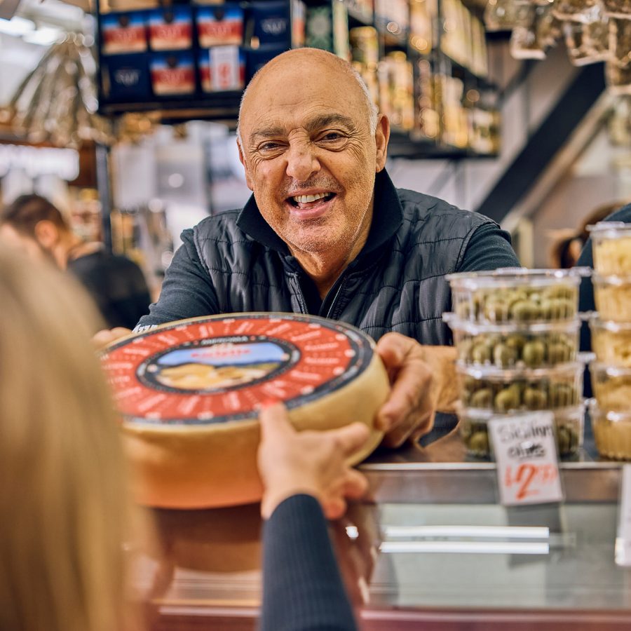 A vendor inside Adelaide Central Market hands a wheel of cheese to a customer. 