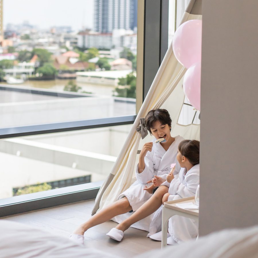 Two young children eating ice cream in bathrobes under a tent by the floor-to-ceiling windows of a family suite in Four Seasons Hotel Bangkok at Chao Praya River.