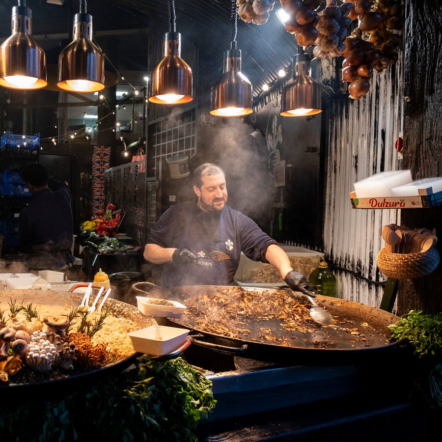 A chef in a black chef’s jacket stirs a big pan of steaming paella in a restaurant.