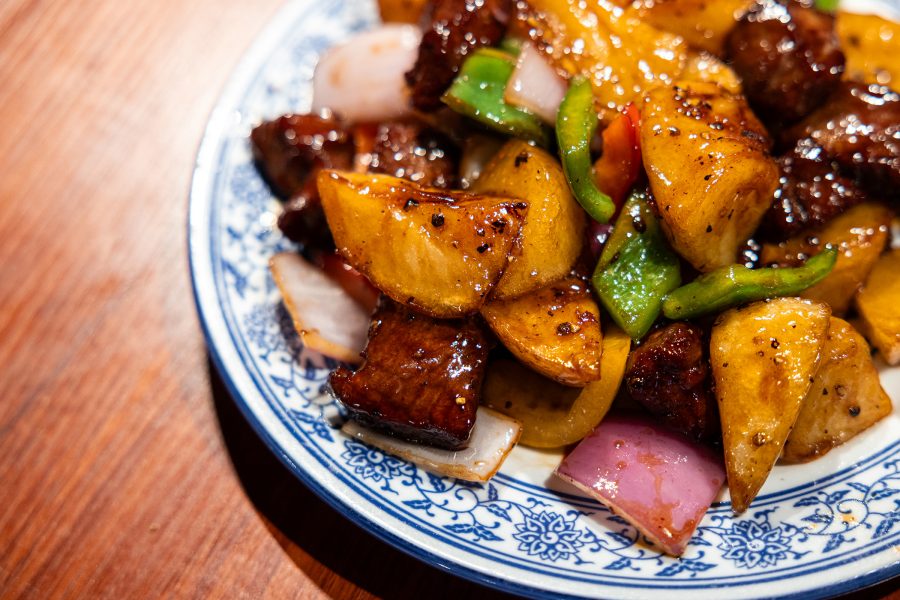 A plate of stir-fried potato and beef cubes with black pepper is placed on a wooden table.