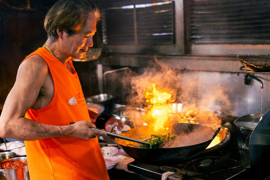 A male chef clad in an orange vest stir fries a dish in a wok bathed in flame.