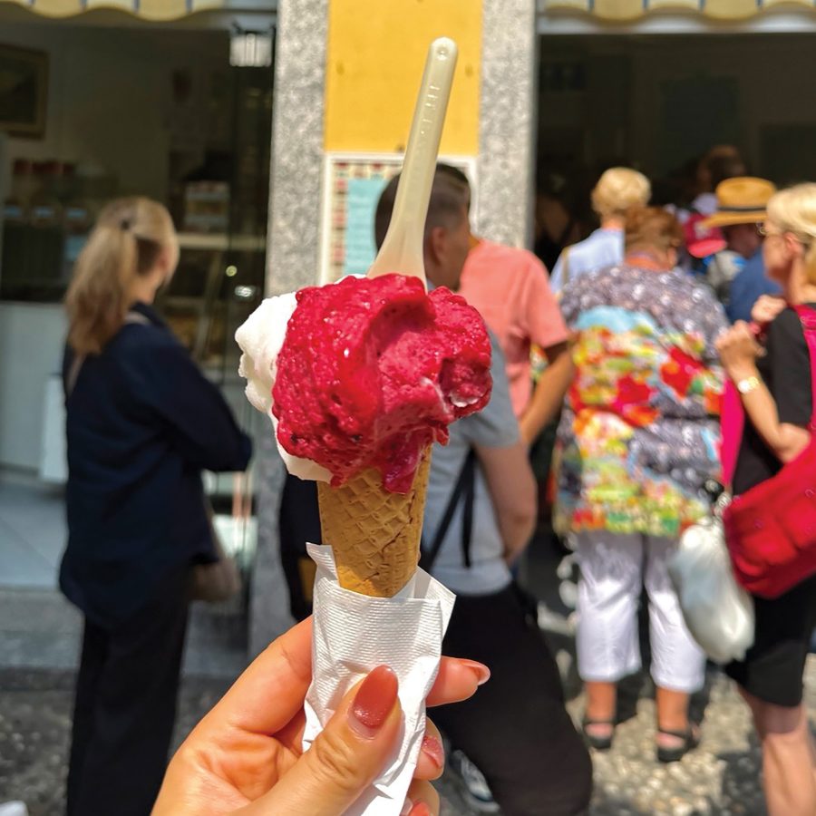 A hand holding up a pink and white gelato in Bellagio, Italy with people in queue in the backdrop