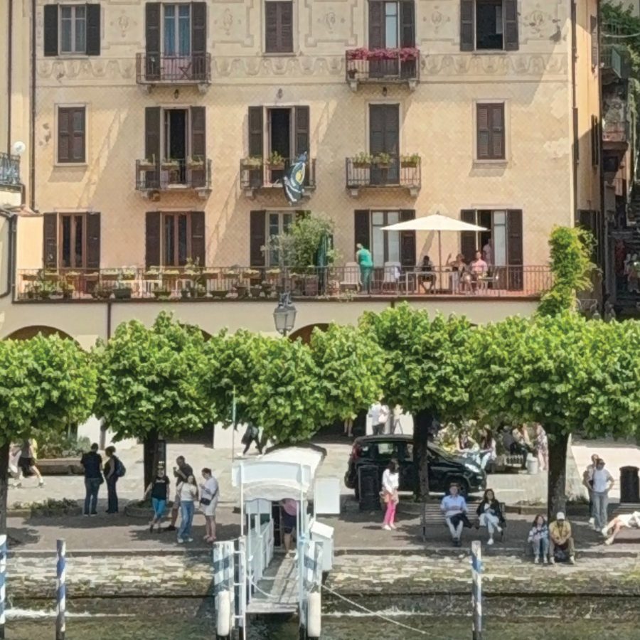 People sitting by the pier under verdant green trees on a sunny day with a view of buildings in the backdrop in Bellagio, Italy