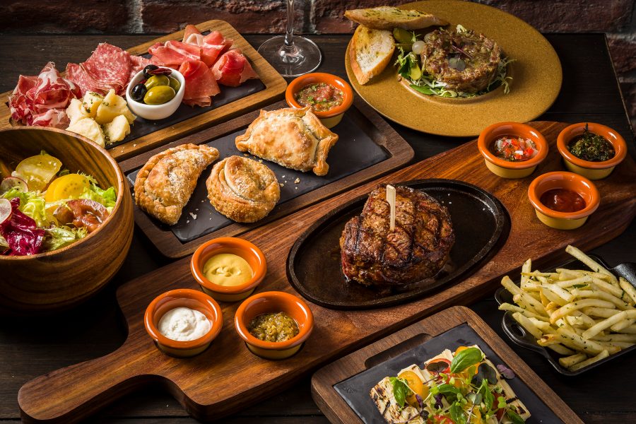 A family spread of various dishes on wooden boards, including steaks, fries, salads, cold cuts and pies, served at Tango Argentinian Steakhouse in Hong Kong 