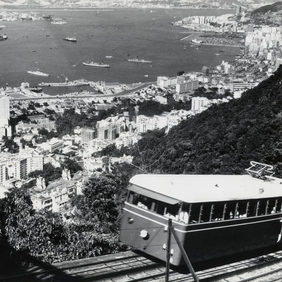 A black and white photo shows the Peak Tram climbing up the mountain as the city of Hong Kong sprawls around the water down below.