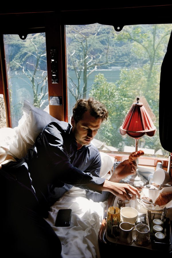 A man leaning on a bed enjoys breakfast foods on his bedside table on board Orient Express La Dolce Vita.