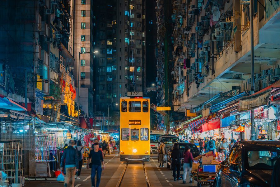 A yellow ding ding tram running down the middle of a street market at night as people walk by in North Point, Hong Kong