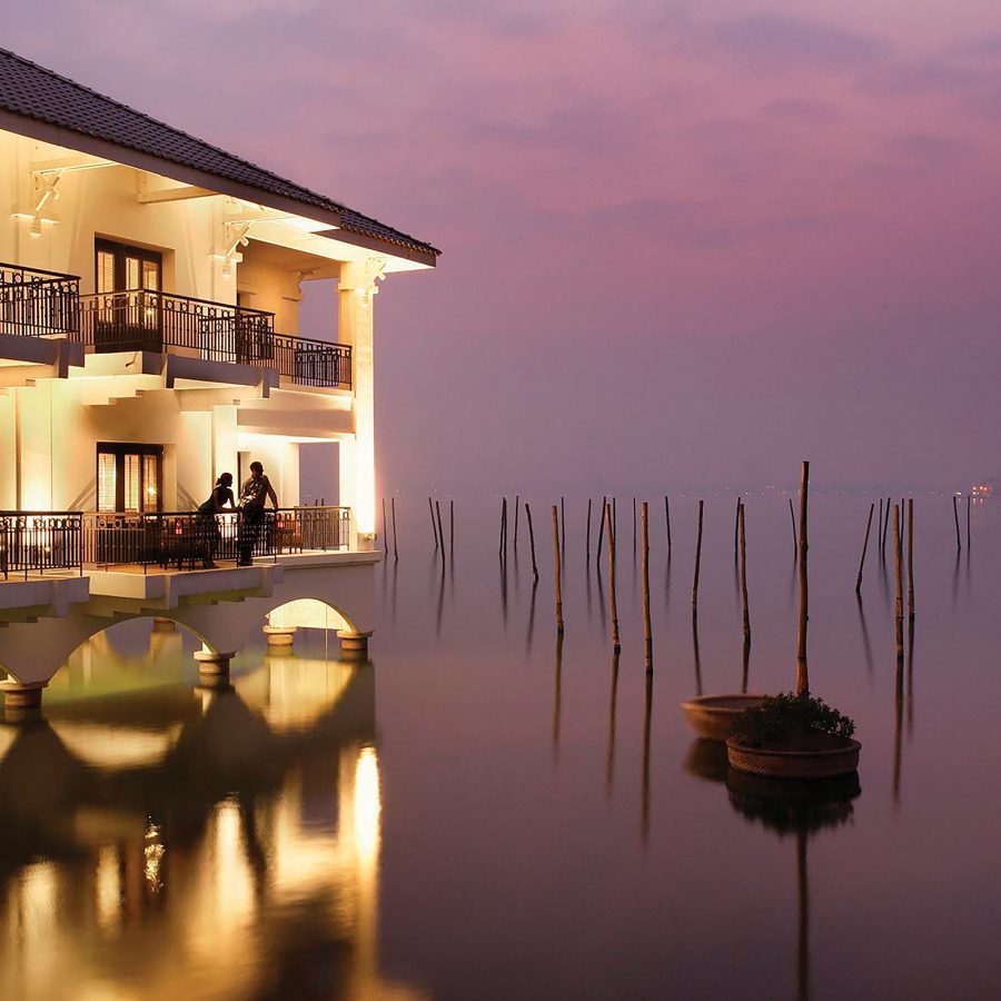 An exterior view of brightly lit InterContinental Hanoi Westlake by IHG in Vetnam after sunset; two people standing on the bedroom balcony overlooking with a view of the lake.