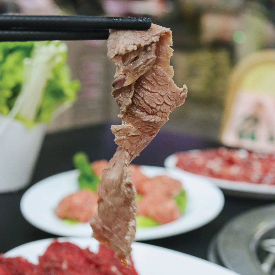 Chopsticks holding up a slice of beef. Plates of food are on the table in the background.