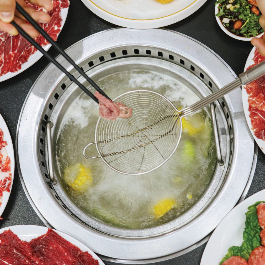 A hand placing a piece of meat into a hotpot using chopsticks. Plates of food are placed around the hotpot.