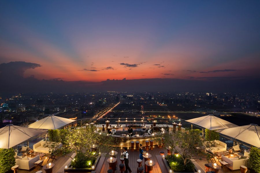 Sunset on a clear night seen from Sora’s sky deck, with the city light of Phnom Penh seen beyond a glamorous outdoor bar.