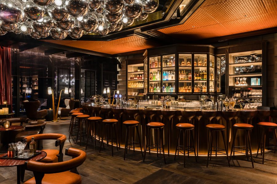 A low-lit curved bar lined with red-topped bar stools and a sculptural ceiling decoration of clustered silver baubles.