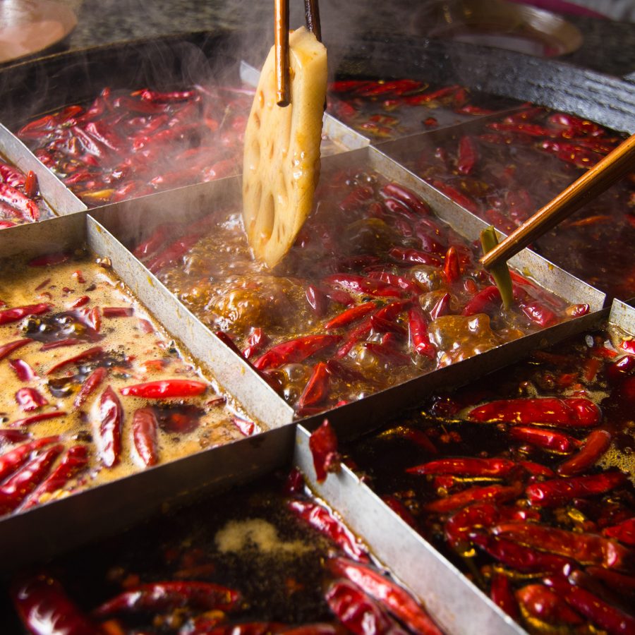 A closeup of a hotpot filled with chilies and a chopstick dipping a slice of lotus root