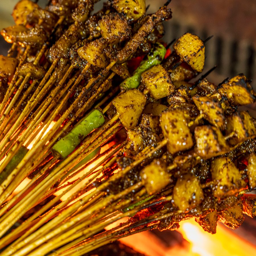 A closeup of a bunch of grilled meats and vegetables on skewers over an open flame 