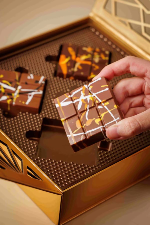 Close-up product image of Dalloyau’s DA CHOCO Golden Autumn Crispy Chocolate, showing a hand holding a piece of chocolate. 