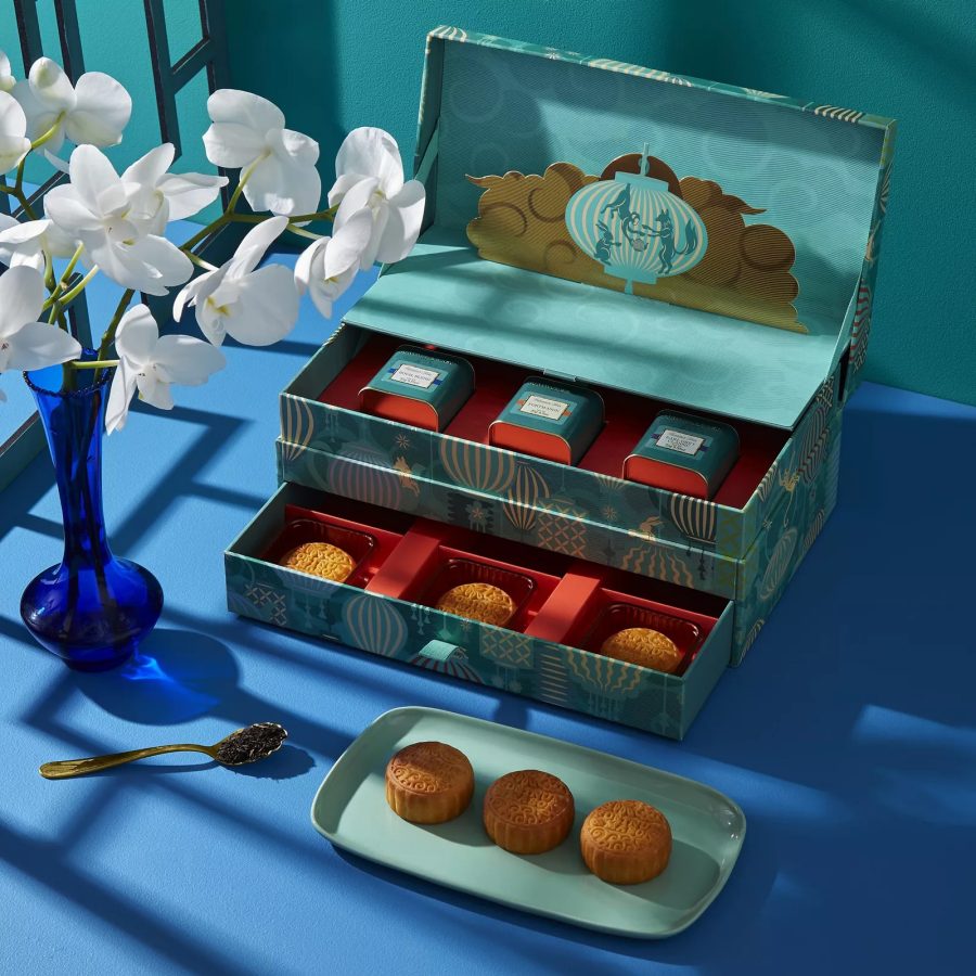 Product image of Fortnum & Mason’s 6-Piece Mooncake & Tea Pairing Giftbox, with three mooncakes placed on a plate in front of a two-tier box with three mooncakes and three tea caddies, next to a spoon and a vase with flowers. 