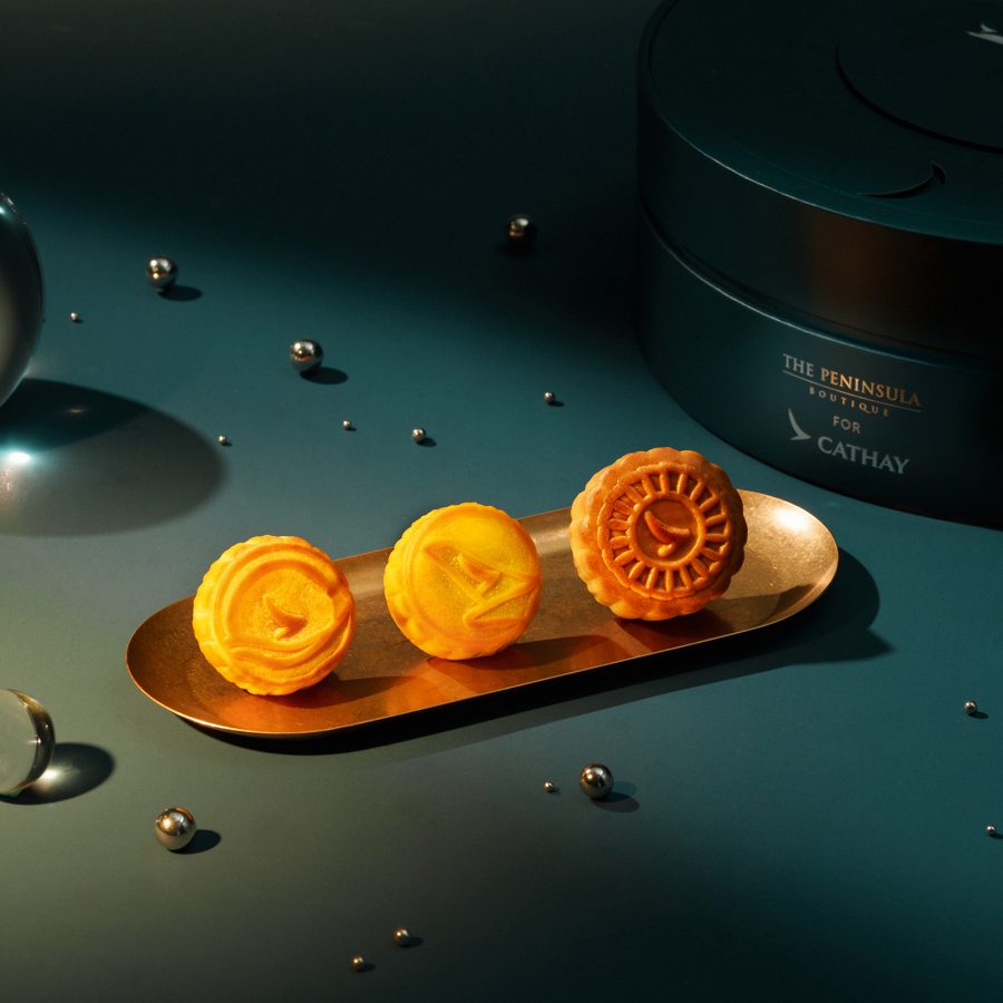 Product image of the Cathay x The Peninsula Boutique Assorted Mini Mooncakes, with three mooncakes placed on a gold plate in front of the round keepsake box. 