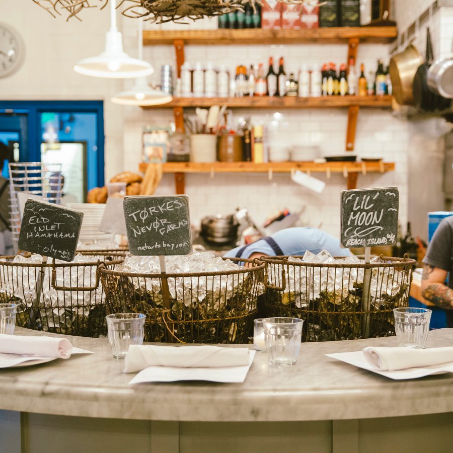 Woman shucks oysters behind the bar at The Walrus and the Carpenter beside iron baskets loaded with shells and ice. 