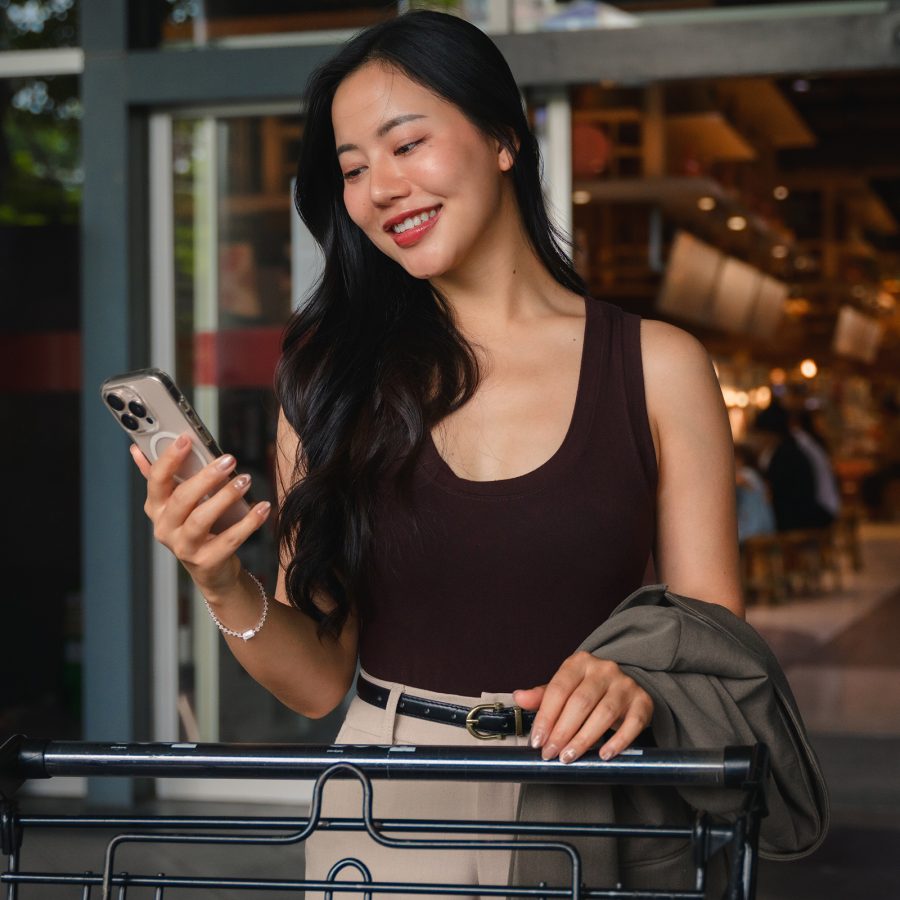 A woman dressed in smart casual outfit smiling while looking at a smartphone in front of a building window.