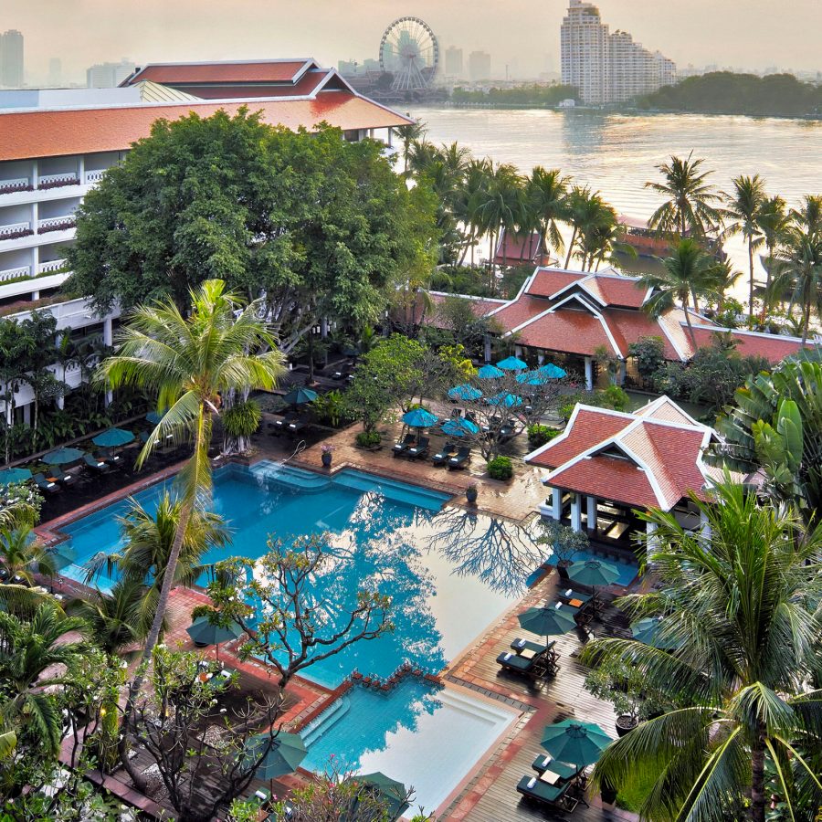 An aerial view of Anantara Riverside Bangkok Resort shows an outdoor pool beneath a large building surrounded by palm trees as the city of Bangkok is seen at a distance. 