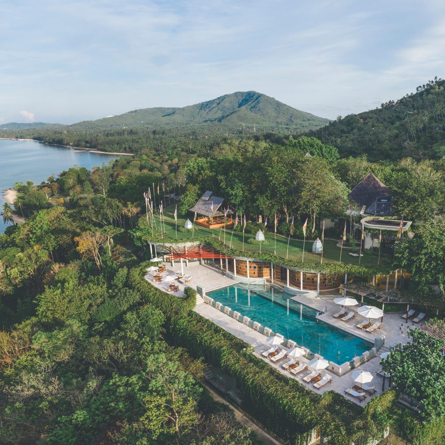 An aerial view of Kamalaya Koh Samui shows a large outdoor pool built into the slope of a tropical forest overlooking the sea.