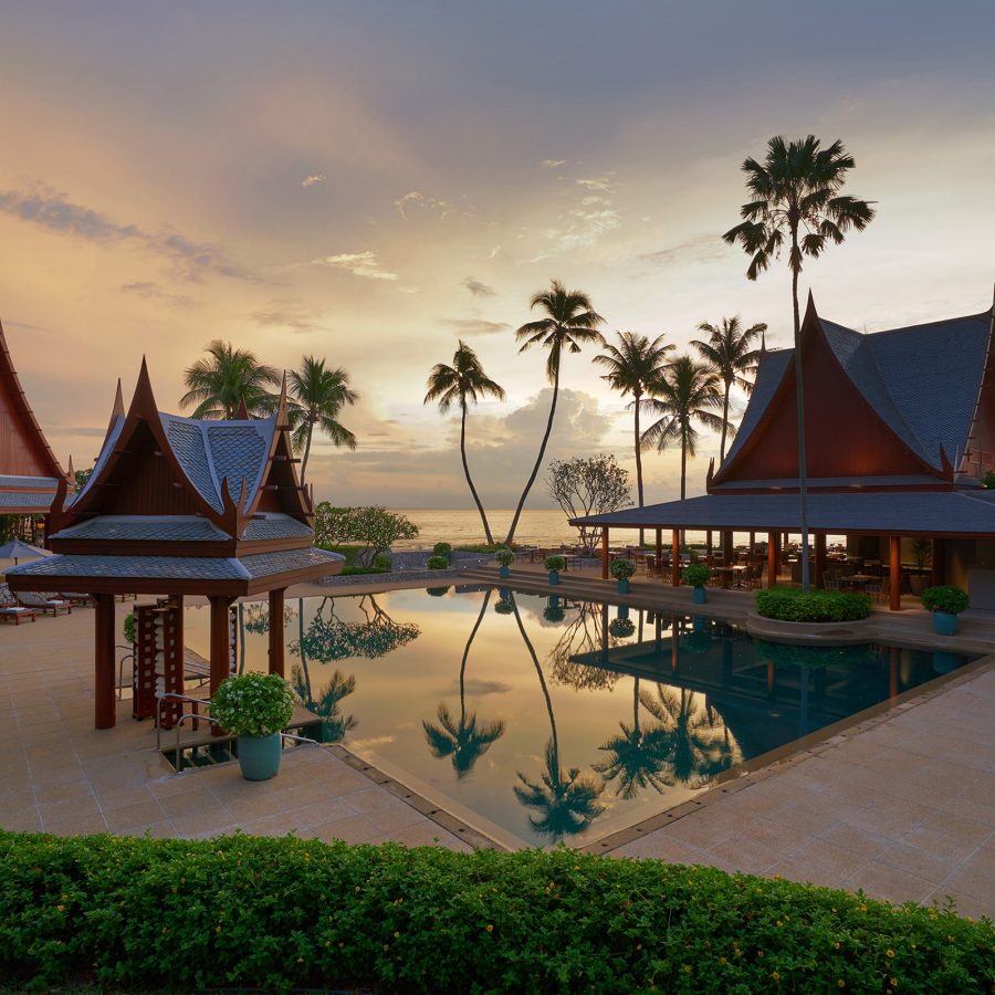 The exterior of Chiva Som Hua Hin is composed of buildings with steep roofs surrounding a pond and set against a wide sky.