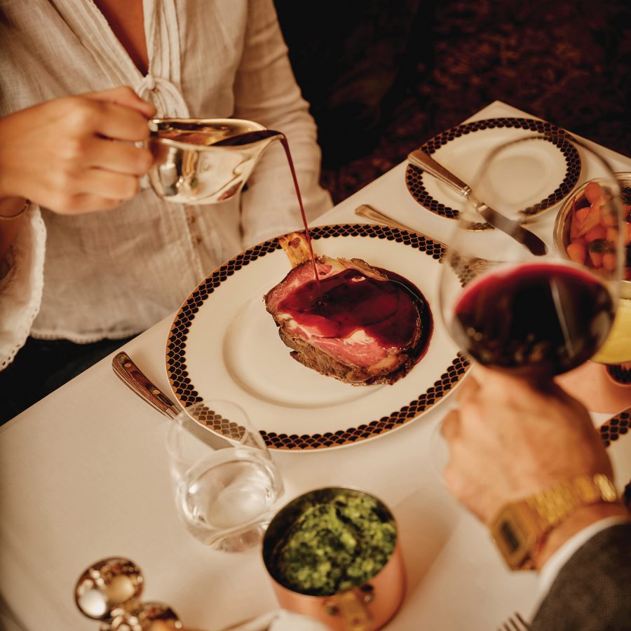 Person pouring gravy over a piece of meat alongside a man holding a glass of wine at The Ned.