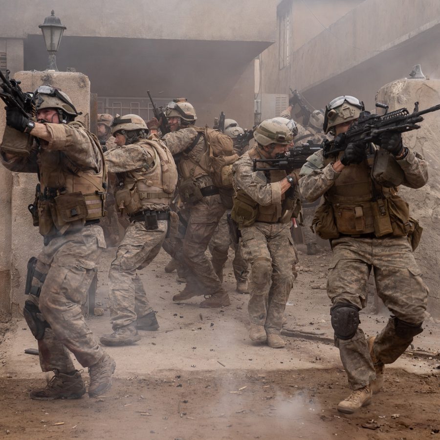 A group of armed force men fire weapons outside a rundown building in a scene from Warfare. 
