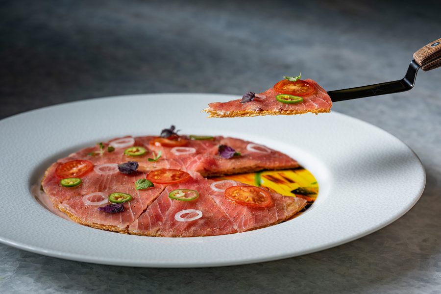 A slice of a flatbread covered in meat is lifted with a knife.