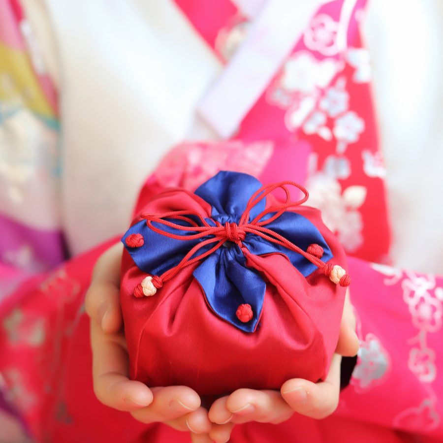 A woman dressed in a hanbok presents a small, wrapped gift.