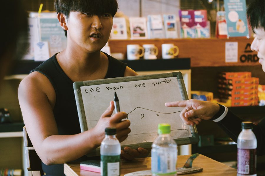 Surf Holic instructor Gyutae Park teaching a class, holding a marker in front of a surfboard that says, “one wave one surfer.” 