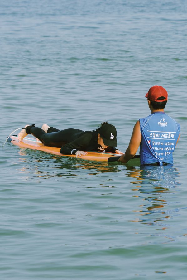 A student lies on a surfboard, which Surf Holic instructor Gyutae Park holds onto while waiting for the next wave.
