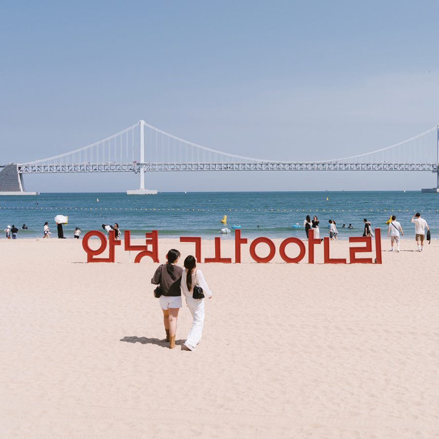 People walking on Gwangalli Beach, in front of an installation on the sand that spells “Hello, Gwangalli” in Korean, and the Gwangan Bridge in the background.