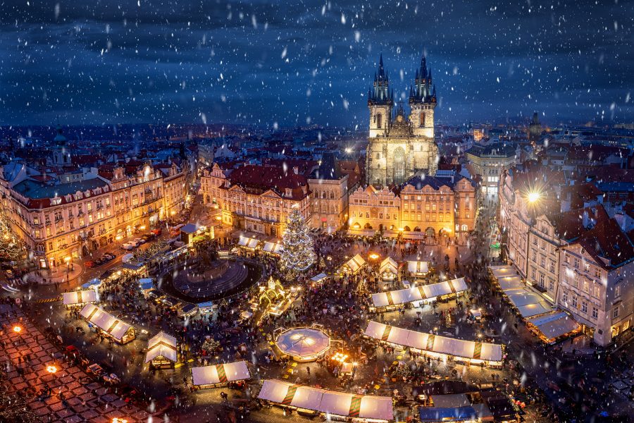 A Christmas market with warmly lit stalls, a large decorated Christmas tree, gently falling snow, and a Gothic church in Old Town Square, Prague. 