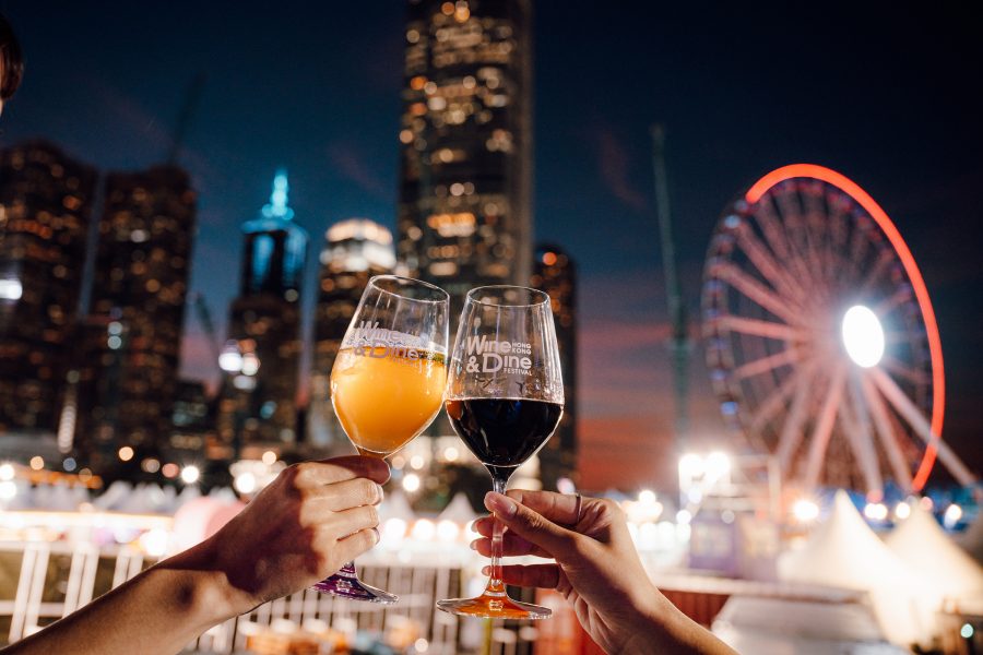 Two people hold up glasses of wine with the Wine & Dine Festival logo on them against a backdrop of the Hong Kong skyline.