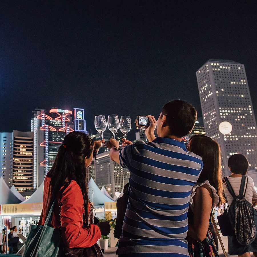 People hold up wine glasses as they take photos of them against the Hong Kong skyline.  