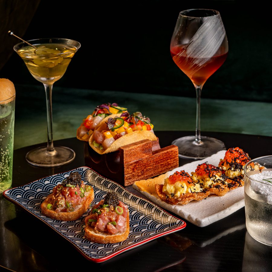 A dark table holds a selection of small tapas-style dishes and bright mixed drinks.