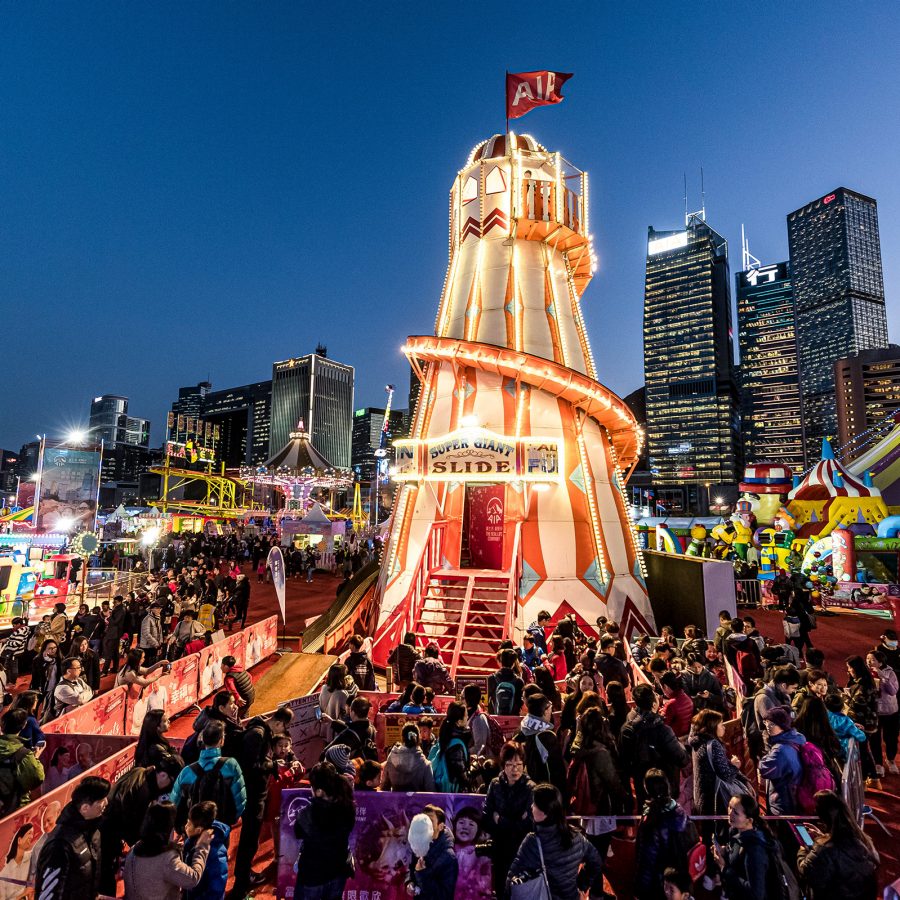 Large outdoor carnival at dusk featuring a brightly lit helter-skelter slide as the centerpiece, surrounded by colourful tents, amusement rides, and a crowd of visitors, with Hong Kong skyscrapers in the background.