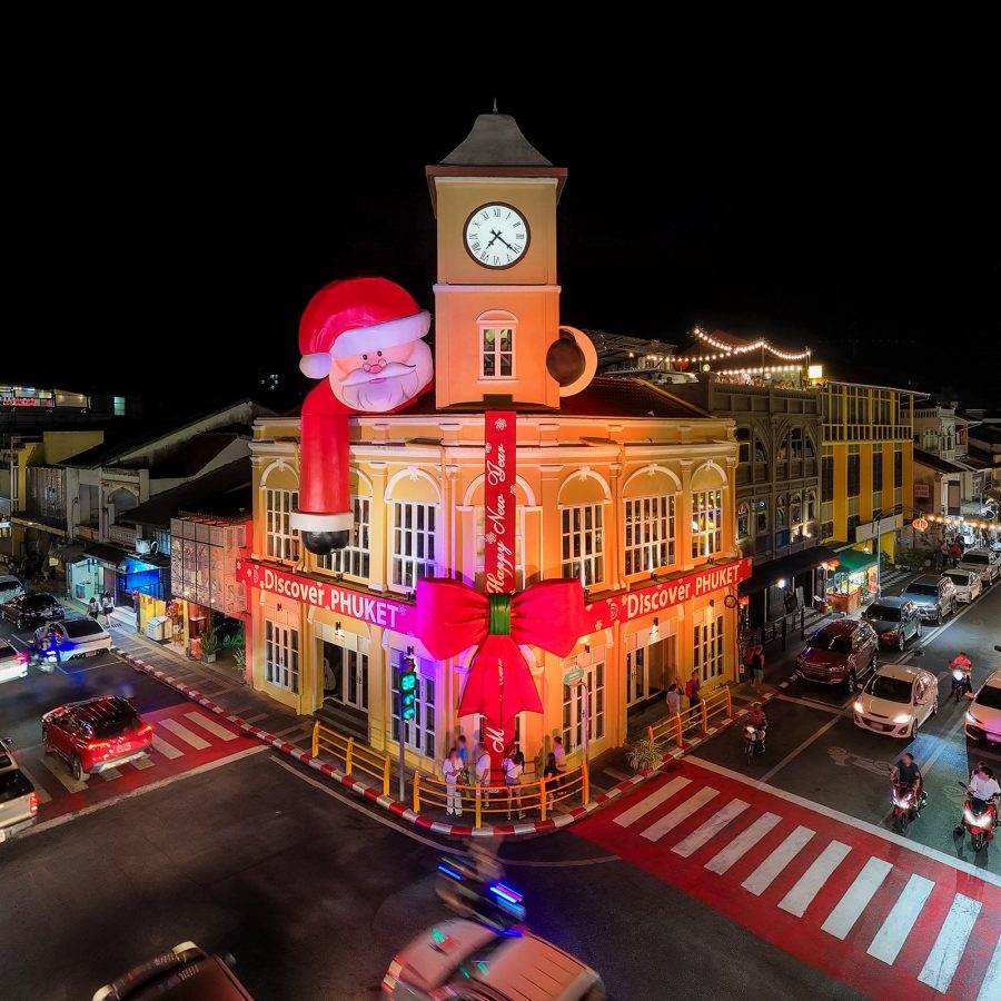 Clock tower in Phuket illuminated with colourful Christmas lights at night, surrounded by festive decorations and street activity.