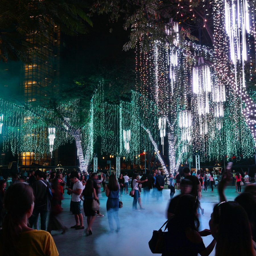 Outdoor light festival at night with trees draped in cascading strings of white and green lights, and a crowd of people walking beneath the illuminated display.