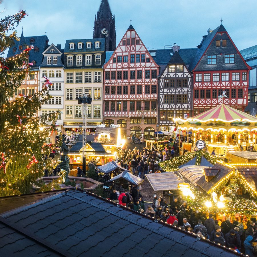 Christmas market in a European square with a large decorated tree, illuminated stalls, and traditional half-timbered buildings.