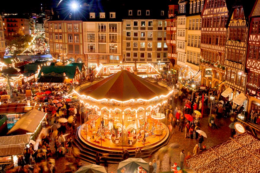 Christmas market at night with a lit carousel, festive stalls, and crowds in a European town square.