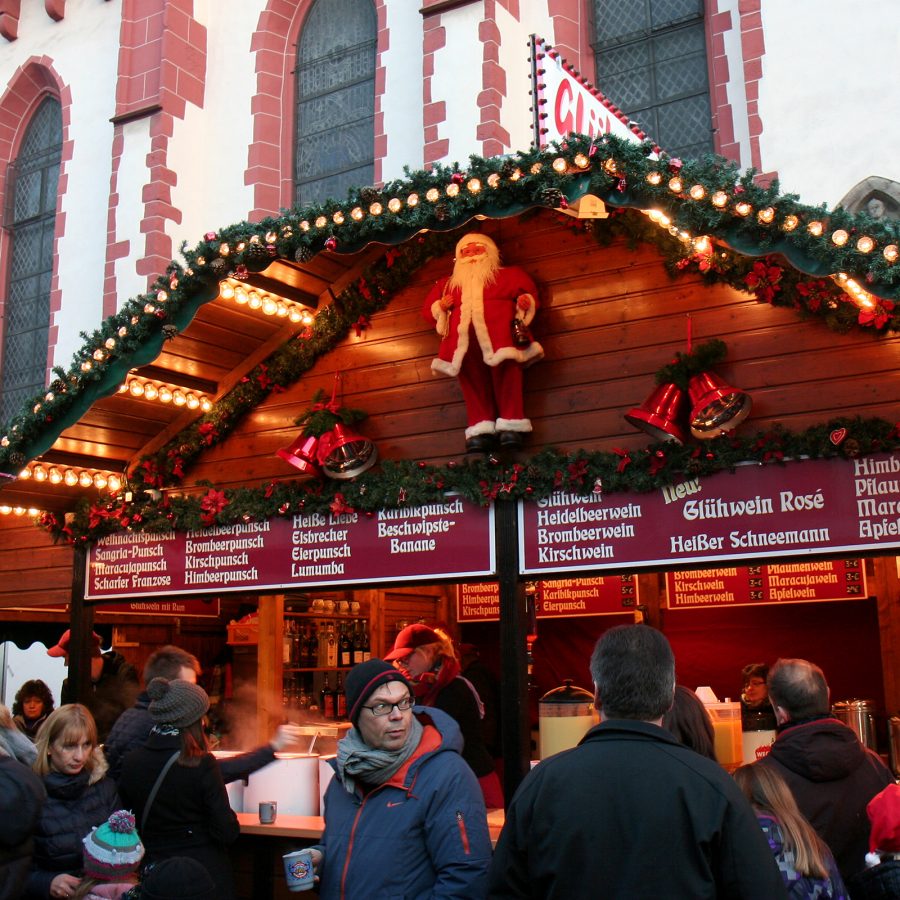 Festive wooden stall at a Christmas market with garlands, lights, and a Santa figure above the counter, surrounded by visitors.
