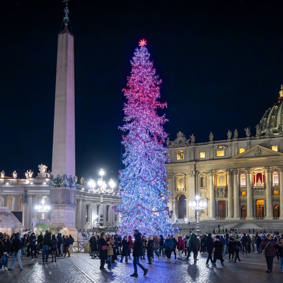 Large Christmas tree with bright lights in front of a historic building in Rome’s city square, with people gathered around.