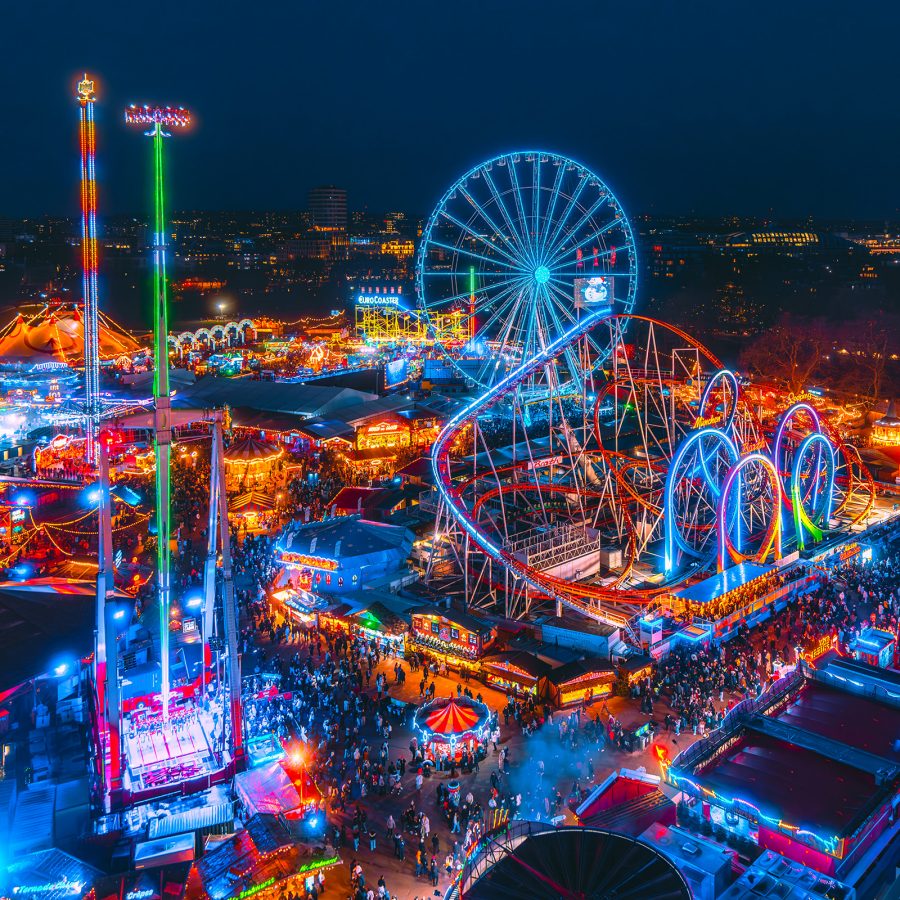 Aerial view of a brightly lit amusement park at night with a Ferris wheel, roller coaster, and colorful rides during a winter festival.