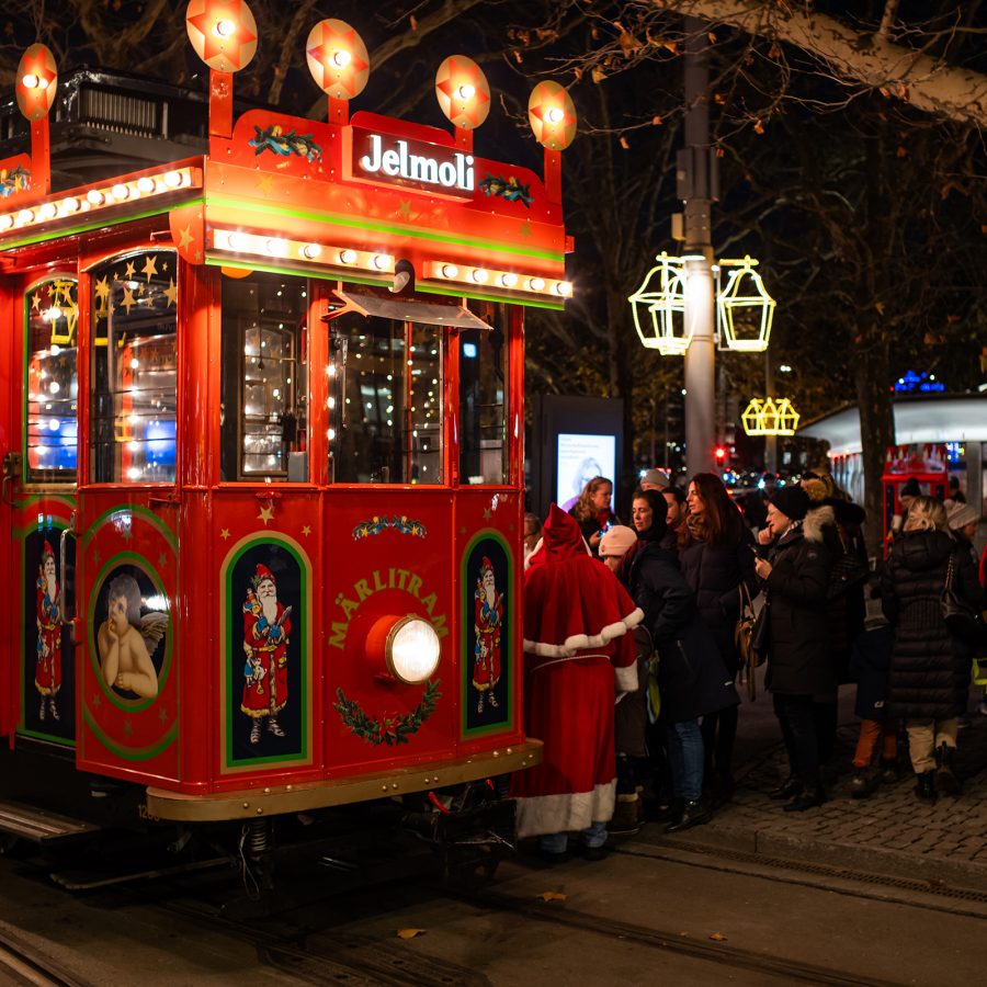 Bright red Christmas tram decorated with festive lights and artwork, parked on a street at night with illuminated trees and a group of people gathered nearby.