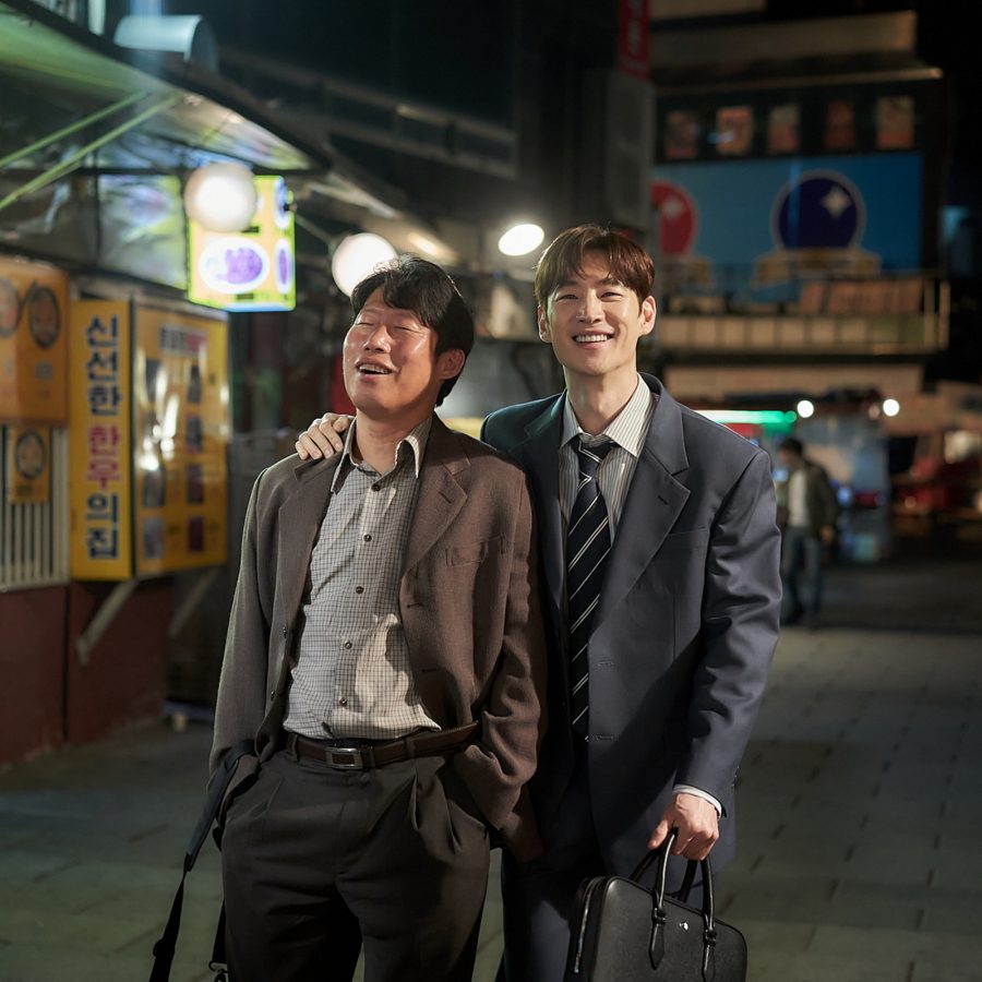 Two smiling men in suits walk down an urban street at night, in a scene from Big Deal
