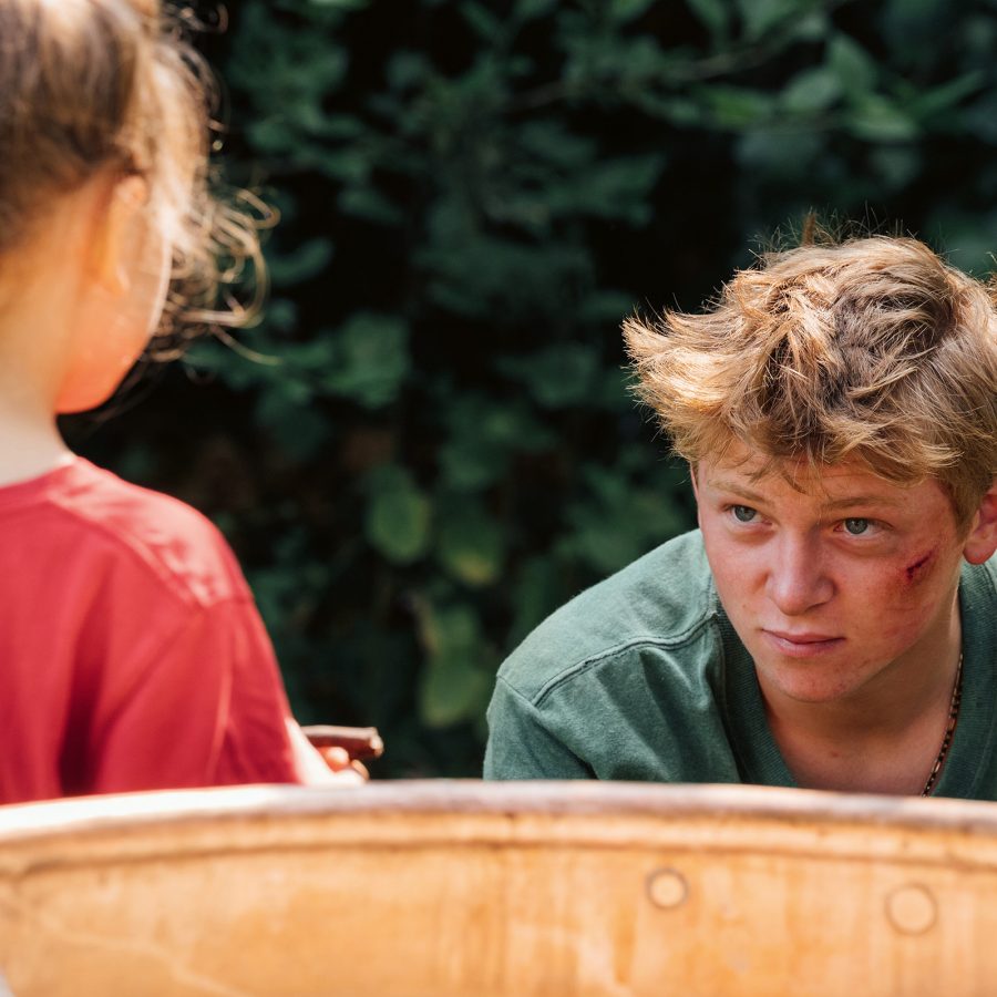 A scruffy-looking teenage boy stands in a garden with a young girl in a scene from Holy Cow