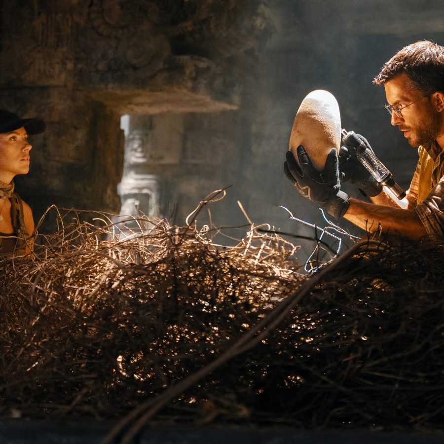 In a scene from Jurassic World: Rebirth, a woman and man stand in a dimly lit ancient building. The man is examining a large dinosaur egg. 
